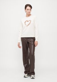 Hvit sweatshirt med hjerte-design i metallisk kobber, kombinert med blanke brune læraktige bukser og mønstrede joggesko.