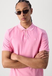 Herre rosa polo skjorte med korte ermer, klassisk krage, plasseringsknapper med tre knapper, og brodert logo på brystet. Brukt med mørke solbriller.