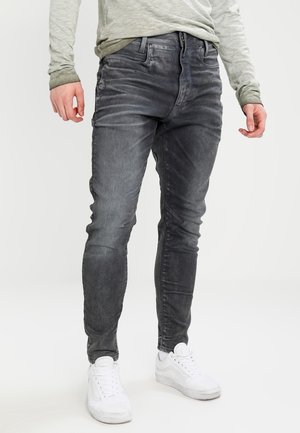 Man draagt strakvallende, vervaagde zwarte jeans, lichtgrijs shirt met lange mouwen en witte sneakers, staand tegen een witte achtergrond.