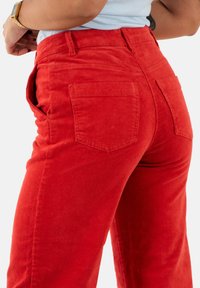 Pantalon en velours rouge, avec des jambes larges, un côtelé vertical et deux poches arrière. Le tissu a une finition douce et texturée.