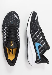 Scarpe atletiche in bianco e nero con una swoosh blu, che presentano un design testurizzato e una suola bianca, poggiate su uno sfondo bianco.