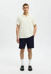Kortärmad, strukturerad polo skjorta i ljus krämfärg med krage, parat med mörk marina shorts, vita strumpor och svarta loafers.