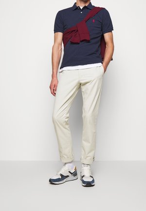 Man in een marineblauwe poloshirt, crèmekleurige broek, marineblauwe en witte sneakers, met een bordeauxrode trui over zijn schouders, staande tegen een witte muur.