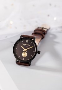 Montre avec bracelet en cuir brun, cadran rond noir, accents dorés et chiffre romain XII. Présente un sous-cadran plus petit à six heures.