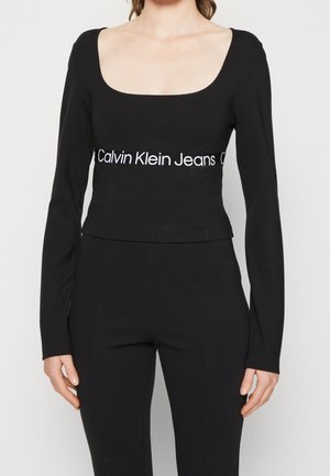 Kobieta ubrana w czarny, długi top odsłaniający brzuch z napisem "Calvin Klein Jeans" pod biustem oraz dopasowane czarne spodnie z wysokim stanem.