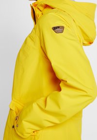 Żółta wodoodporna kurtka z kapturem, kieszenią boczną i naszywką z logo na górnej części ramienia, przedstawiona z boku na osobie.