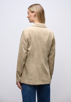 Vrouw in een beige corduroy blazer en blauwe spijkerbroek, staand met haar rug naar de camera tegen een effen lichte achtergrond.