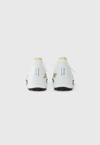 Witte sportschoenen met een zwarte zool, voorzien van een textuurontwerp, zwarte en gouden accenten, en veters. De achteraanzicht toont de logo-details.