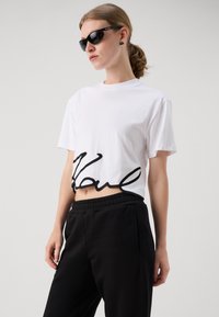 Balta trumpa T-shirt su juodu siuviu "Karl" ant apatinio krašto. derinama su juodomis elastingomis kelnėmis. Glotni audinio tekstūra.
