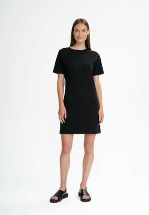 Schwarzes T-Shirt-Kleid, kurze Ärmel, gerader Schnitt. Glatte Textur mit schlichtem Design, kombiniert mit flachen schwarzen Sandalen.