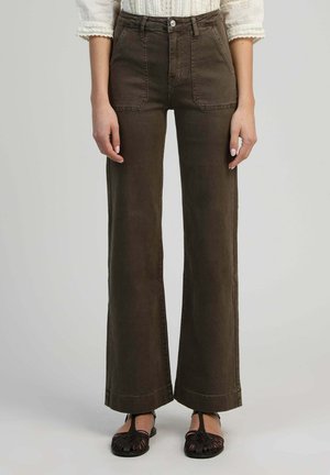 Femme portant un pantalon large taille haute marron foncé avec poches avant, associé à un chemisier blanc à poignets en dentelle et des sandales noires tissées.