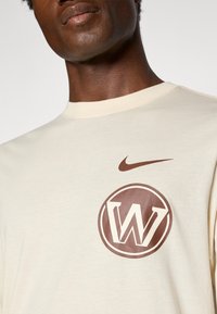 Kermanvärinen puuvillapaita, jossa on pyöreä ruskea logo, jossa on tyylitelty "W" ja ruskea Nike-swoosh vasemmalla olkapäällä.