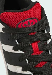 Black and white sneaker with red mesh tongue, featuring black laces and a glossy red logo, textured upper, and distinct three stripes design.