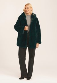 Faux fur green coat with a wide collar and loose sleeves, paired with black flared pants and a light gray ribbed top.