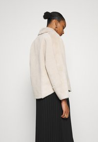 Beige faux fur jacket with a soft texture, wide sleeves, and a collar. Features rounded hem and a relaxed fit, worn over a pleated black skirt.