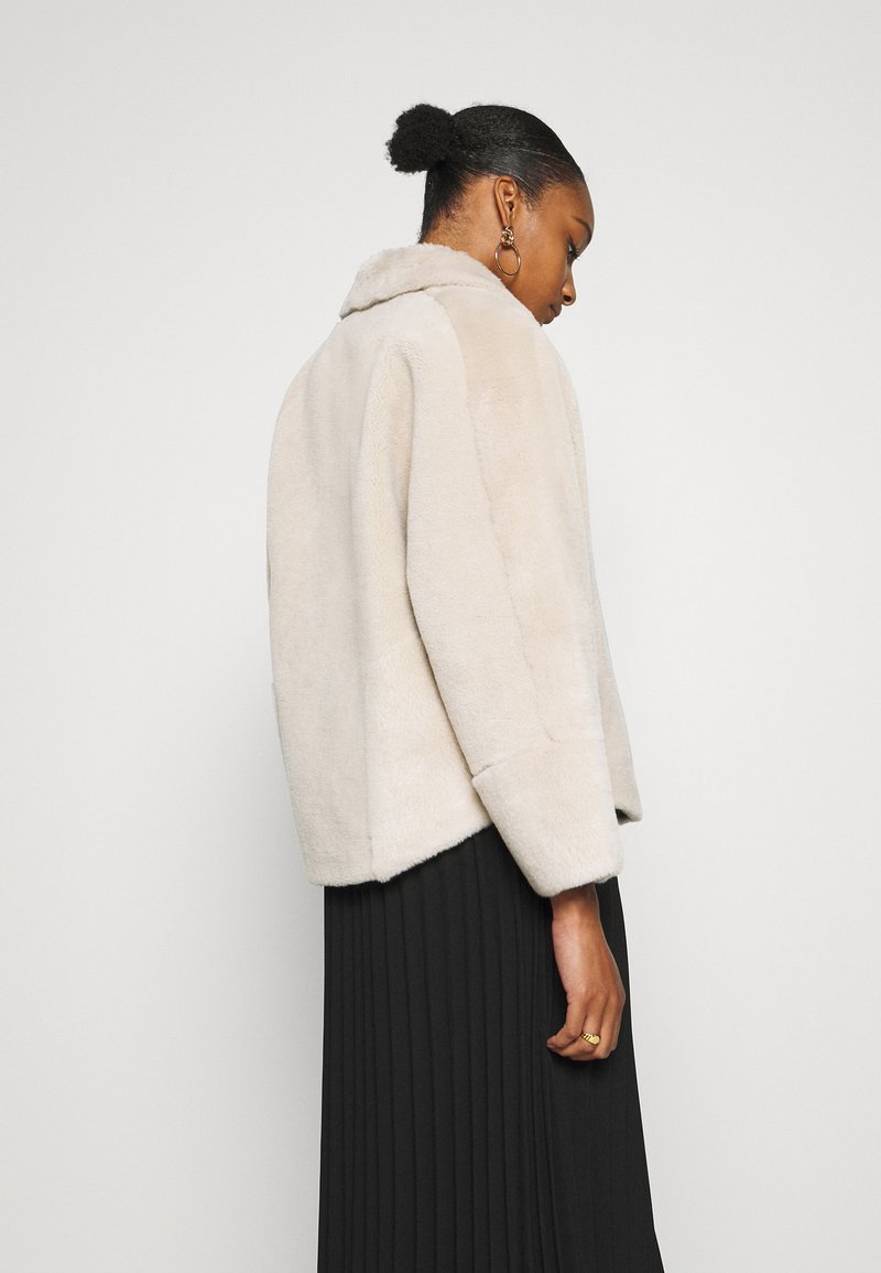Veste en fausse fourrure beige avec une texture douce, des manches larges et un col. Caractéristiques : ourlet arrondi et coupe décontractée, portée sur une jupe noire plissée.