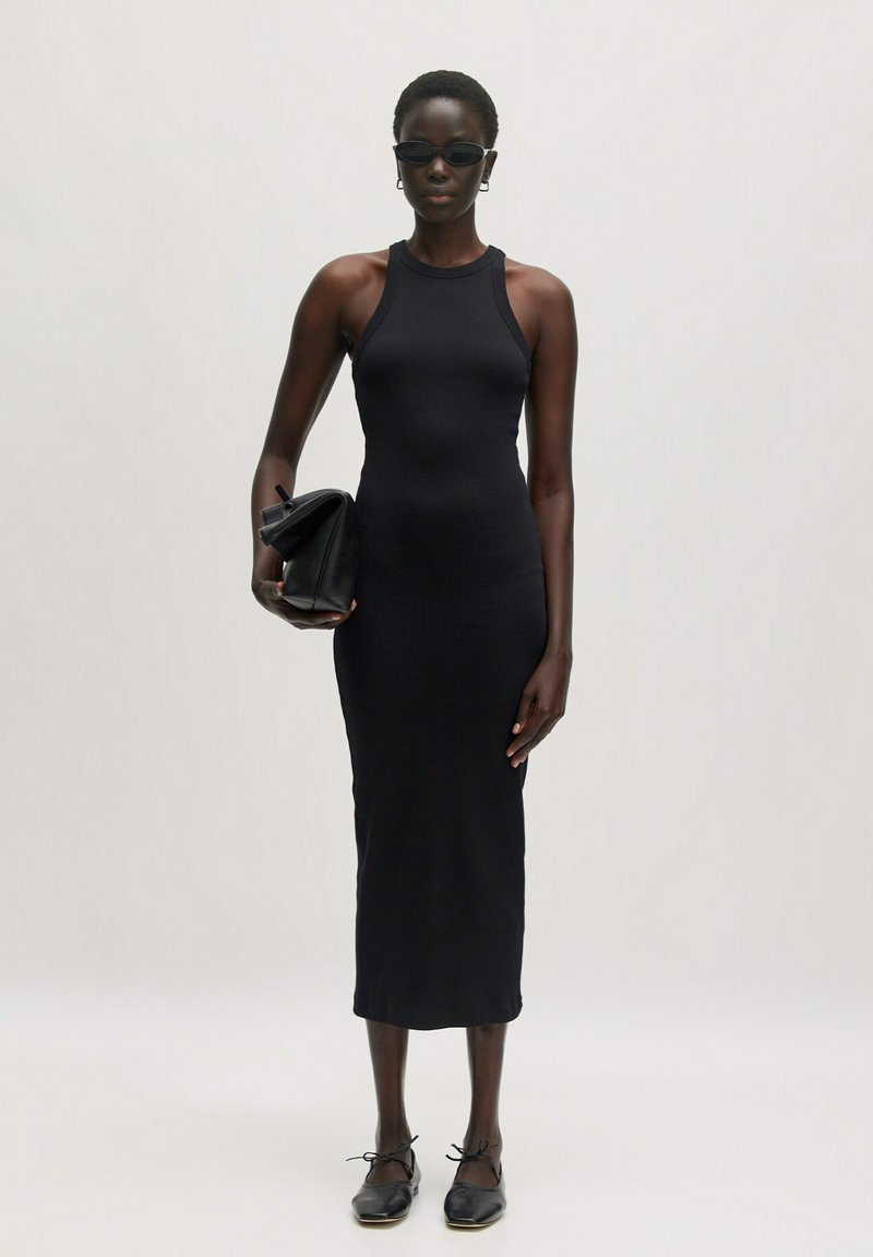 Schwarzes, figurbetontes, geripptes Midikleid mit hohem Hals. Kombiniert mit einer schwarzen Clutch und flachen schwarzen Schuhen, die eine elegante Silhouette schaffen.