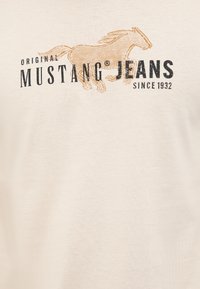 Cream-colored t-shirt featuring the text "Original Mustang Jeans" in black with a running horse graphic in gold. Cotton fabric, short-sleeved.