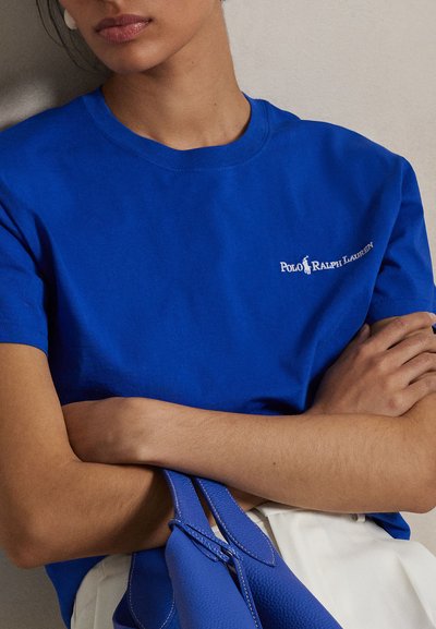 Sieviete ar sakrustotām rokām, valkājot zilu Polo Ralph Lauren t-kreklu un turot roku zilu somu uz neitrālas krāsas fona.