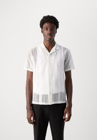 Cămașă albă cu mânecă scurtă, cu modele delicate din dantelă, cu nasturi pe față și tiv drept. Purtați-o cu pantaloni negri.
