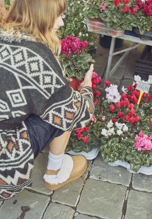 Beige slipper with white fur accents, worn with white socks. Individual is crouching beside pots of vibrant pink and white cyclamen flowers.