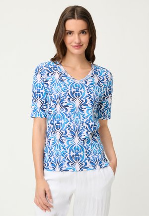 Kobieta nosząca niebiesko-biały wzorzysty top z krótkim rękawem i dekoltem w serek oraz białe spodnie, stojąca z jedną ręką w kieszeni, uśmiechająca się.