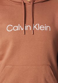 Brun hættejakke med et hvidt "Calvin Klein" logo. Lavet af blød bomuld, har den en frontlomme og justerbar snørelukning.