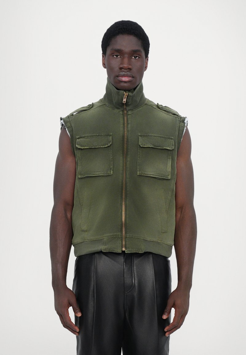 Gilet sans manches vert olive avec col haut, fermeture éclair à l'avant, poches poitrine doubles et coupe décontractée, associé à un pantalon en cuir noir.