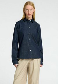 Tamsiai mėlynas džinsinis marškinis su apykakle, ilgomis rankovėmis ir baltais mygtukais; turi tiesų siluetą ir minimalistinį dizainą.