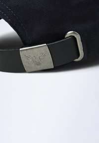 Casquette en tissu marine avec une sangle en cuir et une boucle en métal, ornée d'un logo ailé gravé. Vue rapprochée de la sangle et de la boucle.