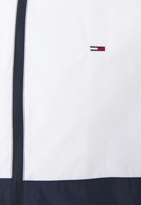 Vit och marinblå vindjacka med frampaneldragkedja, prydd med en liten broderad logga. Slätt och lätt tyg med kontrasterande sömmar.