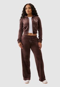 Kobieta stoi w brązowej welurowej bluzie z zamkiem, dopasowanych spodniach, białym topie crop i białych sneakersach na jednolitym szarym tle.