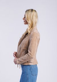 Blonde vrouw in een beige suède jas en blauwe jeans staat in profiel tegen een witte achtergrond, met haar handen gekruist vlak bij haar taille.