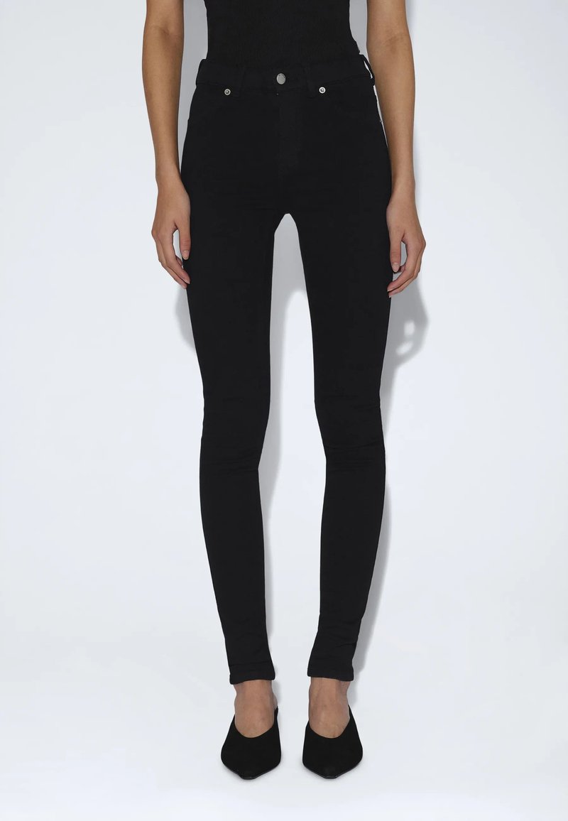 Personne portant un jean skinny noir taille haute et des talons noirs à bout pointu, debout devant un fond blanc uni.