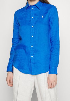 Vrouw draagt een felblauw overhemd met lange mouwen en knopen, met een klein wit logo op de borst, en witte broek.