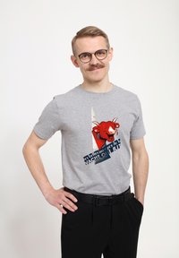 Šedé bavlněné tričko s grafikou červené krávy a textem, kulatý výstřih, krátké rukávy, v kombinaci s černými kalhotami a černým páskem.