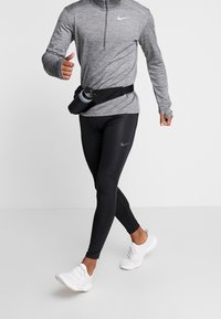 Gray long-sleeve athletic shirt with a half-zip and thumbholes, paired with black leggings and white running shoes; black water bottle on waistband.