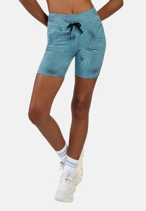 Blaue, gemusterte High-Waist-Fahrradshorts mit schwarzem Kordelzug, getragen mit weißen Knöchelsocken und weißen Sportschuhen an gekreuzten Beinen.