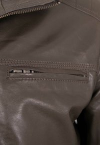 Dark brown leather jacket detailing a zippered pocket and stitching accents. The texture appears smooth with subtle sheen.