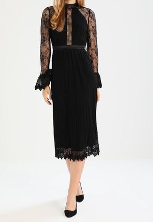 Femme portant une robe midi noire avec des manches longues en dentelle, un col en dentelle transparente, une taille cintrée et un ourlet en dentelle festonnée, assortie à des talons noirs.