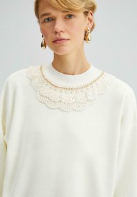 Witte trui met een geribbelde kraag, voorzien van een delicate gehaakte kantdetail rond de halslijn. Model draagt gouden druppel oorbellen.