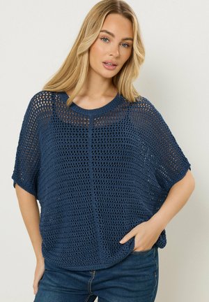 Donna bionda che indossa un maglione lavorato a maglia aperta blu navy sopra una maglietta nera, abbinato a jeans blu scuro, posando con una mano in tasca.