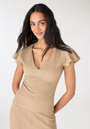 Donna con lunghi capelli scuri che indossa un vestito beige a V con maniche svolazzanti e trama struttura, sorridente davanti a uno sfondo chiaro e semplice.