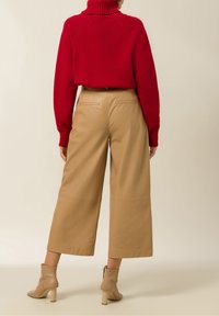 Rode gebreide coltrui gecombineerd met beige wijde faux leren broeken en beige enkellaarsjes met blokhak, gezien vanuit de achterkant.