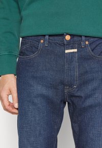 Mörkblå denimjeans med en klassisk rak passform. Har kopparfärgade detaljer, orange sömmar och en liten tygetikett på midjebandet.