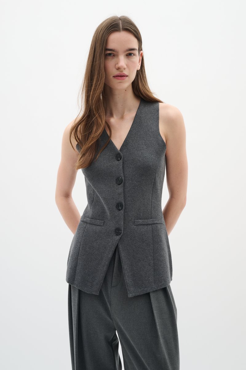 Gilet grigio senza maniche in misto lana, con scollatura a V profonda, cinque bottoni neri e due tasche frontali, abbinato a pantaloni grigi a gamba larga.