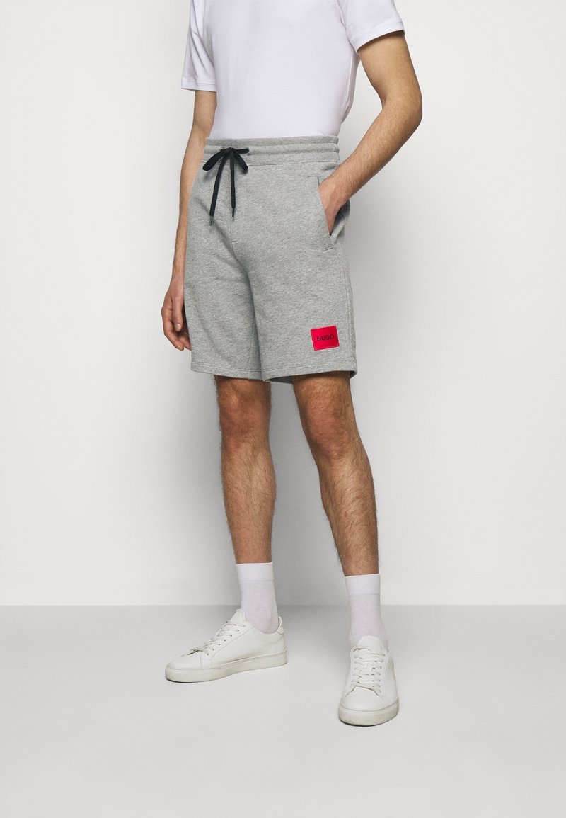 Shorts en sweat gris avec une taille élastique et un cordon noir. Comprend des poches latérales et un patch logo rouge sur la cuisse gauche.