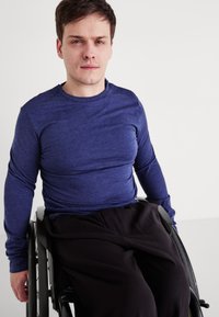 Kék, hosszú ujjú ing puha anyagból, fekete nadrággal párosítva. Az illető egy kerekesszékben ül, bemutatva az alkalmazkodó ruházatot.