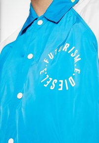 Modrá voděodolná bunda s bílými patentkami. Obsahuje kruhový text "FUTURISM" a "DIESELT" v kontrastním písmu. Hladká textura.