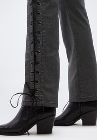 Γκρι τζιν με πλαϊνή λεπτομέρεια κορδονιών και μαύρα ακραία, συνδυασμένα με μαύρα μπότες αστραγάλου με τακούνι, που έχουν λείο δέρμα και μυτερή μύτη.